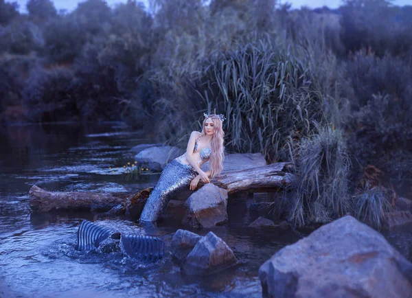 Affascinante sirena snella sdraiata sulle pietre nel fiume acqua serale, una bionda con una corona di mare sulla testa e una lunga coda con squame, crea onde con una pinna, regina del mondo sottomarino — Foto Stock