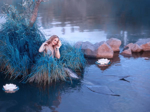 Encantadora menina atraente com cabelo longo onda loira senta-se na grama no meio do lago frio, coloca o cabelo para trás e sorri, segure uma enorme concha perto da orelha e ouvir o som do mar — Fotografia de Stock