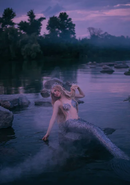 Sirena rubia baja su mano, larga cola de plata deliciosa en el agua, crea niebla con su magia, atractivo mago de mar con tiara descansando sobre piedra en medio de un bosque tranquilo en el río — Foto de Stock