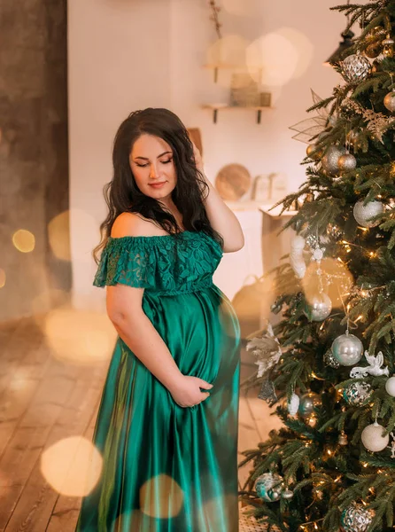 Ganska mörkhårig gravid kvinna i underbara bedårande grön smaragd lång klänning nära nyår träd, lady blygsamt sänkte ögonen ner, sätter håret tillbaka, kramar sin mage, bokeh från julbelysning — Stockfoto
