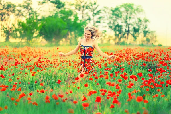 Allegra bella ragazza attraente con i capelli biondi ricci volanti corre attraverso il campo di papavero e ride, il vento soffia sul suo viso, vestito con un abito rosso carino corto con le spalle nude aperte — Foto Stock