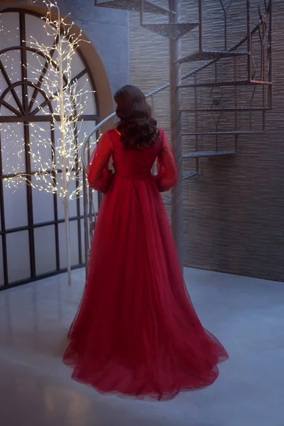 Дівчина в чудовому червоному розкішному довгому платті зі спиною до камери, демонструє темне волосся з ідеальними хвилями, кімнатою в стилі лофт, яскравими один на дереві. Фото без обличчя . — стокове фото
