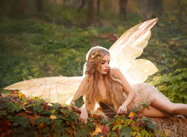 Una dríada delgada, una ninfa con el pelo largo rubio y una corona de oro se encuentra en las hojas en el bosque en un vestido largo beige con las piernas desnudas, tiene alas brillantes detrás de su espalda, gran foto de verano — Foto de Stock