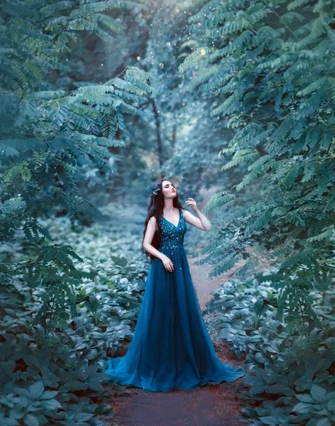 Attractive charming lady with dark hair and white skin like sleeping beauty, lady with closed eyes in middle of emerald forest on fabulous trail in long blue gorgeous brilliant dress with sparkles — Stock Photo, Image