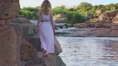 ince uzun boylu model sarışın kıvırcık akan saçlı yavaş yavaş yalınayak taş, bir orman perisi veya nehir peri uçan hem, yüzsüz, parlak sulu renkler ile uzun pembe yaz ışık elbiseli yürür