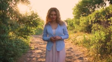 sarışın Bayan yalnız düşünceli bir yeşil yemyeşil orman kumlu bir yoldaki, yaprakları ile parlak güneş ışınları, bir kız uzun pembe bir sundress ve bir hipster gülümsüyor gibi mavi bir gömlek boyunca yürür