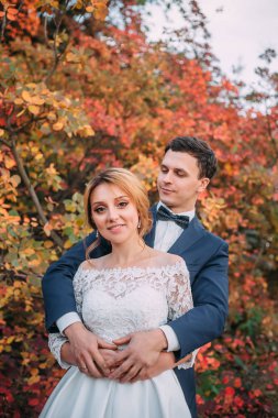 İnanılmaz çekici genç çift düğün günü. zarif beyaz uzun elbise ve mavi buketi Gelin el, damat mavi moda iş takım elbise, kırmızı ağaçlarda ayakta ve sarılma