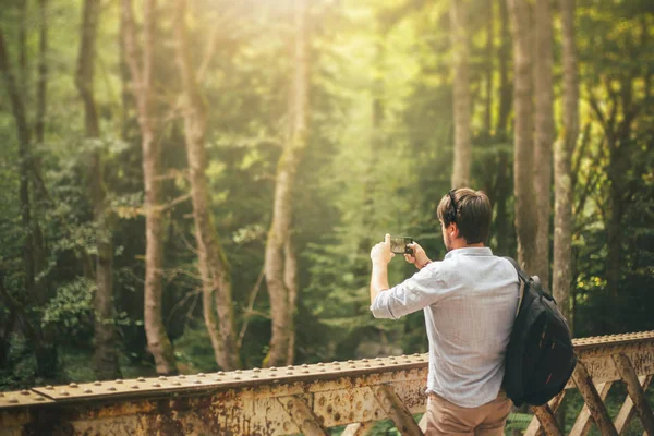 Un hombre moreno en un puente sobre un río y fotografiando la exuberante y vibrante naturaleza en Georgia con un nuevo teléfono. una idea para la publicidad de un gadget y cosas, un turista con una mochila negra — Foto de Stock