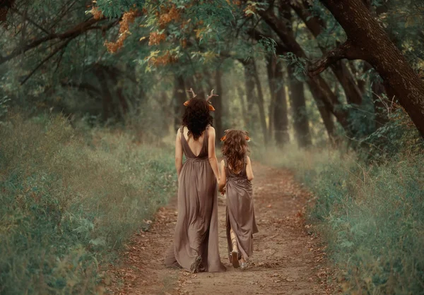 Креативна сімейна фотографія брюнетки мати і блондинка дочка, заводи тримаються за руки і йдуть глибоко в ліс уздовж покинутої таємної доріжки, одягнені в довгі світло-коричневі сукні з вирізами на спині — стокове фото