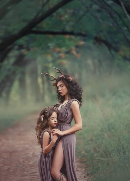 Two girls in the form of deer hug and close their eyes, fauns in long brown dresses, a cutout reveals the mothers bare leg, magical fairytale makeup, homemade wonderful animal horns and ears — Stock Photo, Image