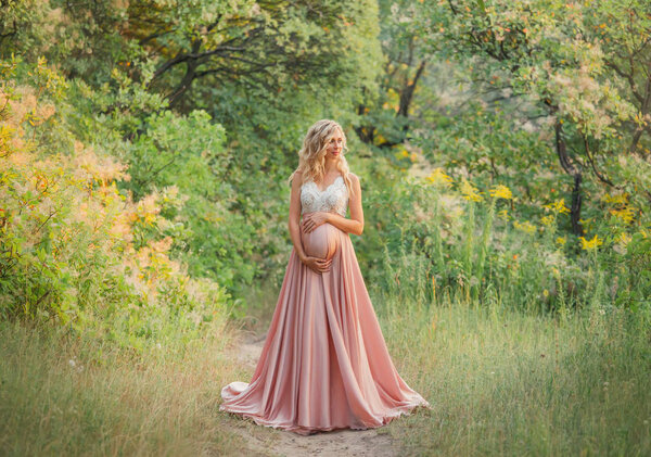 Young sweet pregnant girl with light curly hair hugs and holds her tummy, standing in a fairy forest in cold shades. dresses a long elegant satin silk pink dress with a lace top. art processing photo.