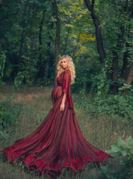 Jeune femme enceinte aux cheveux bouclés blonds en robe écarlate rouge battante longue lumière, debout dans la forêt, tenant, étreignant son ventre. comme une magnifique reine des fées. art automne sombre halloween photo — Photo