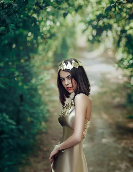 Lyxiga brunett tjej poserar i en gyllene stål klänning och bär en krona med blad. Gothic Queen hösten i mystiska skogen. Prinsessa med brutala ögon tittar på kameran — Stockfoto
