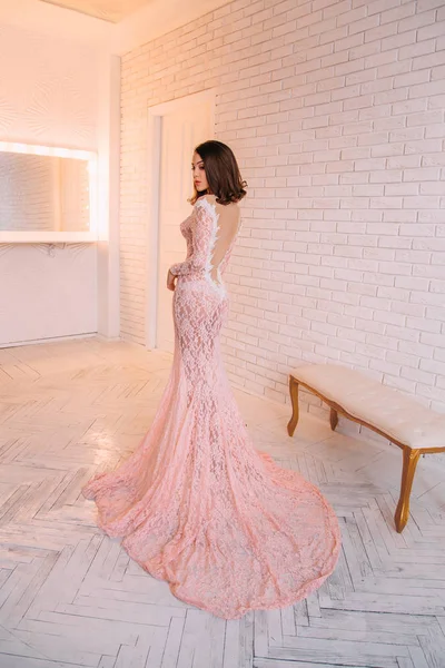 Luxurious brunette girl, with a bob haircut, posing in an expensive, pink dress with an open back. The princess in the white room. Scandinavian minimalist style interior. Image for prom and party
