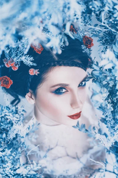 Orgullosa majestuosa reina del invierno y eterno frío en vestido blanco largo con pelo oscuro recogido adornado con rosas congeladas y bayas, una hermosa chica en el reino de hielo, una imagen sombría en colores brillantes —  Fotos de Stock