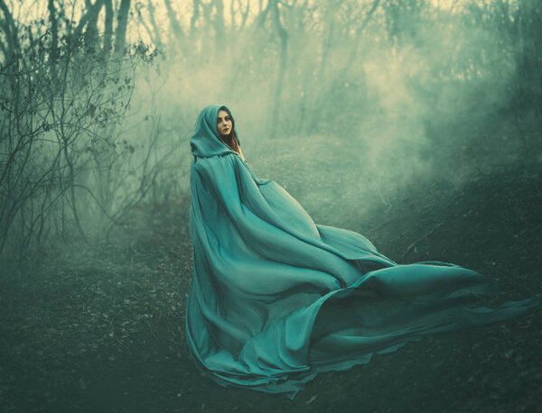 Attractive large lady in long blue summer light waving raincoat runs through forest with bare trees and mysterious white smoke and magical fog, charming sorceress escapes from a fairytale monster.