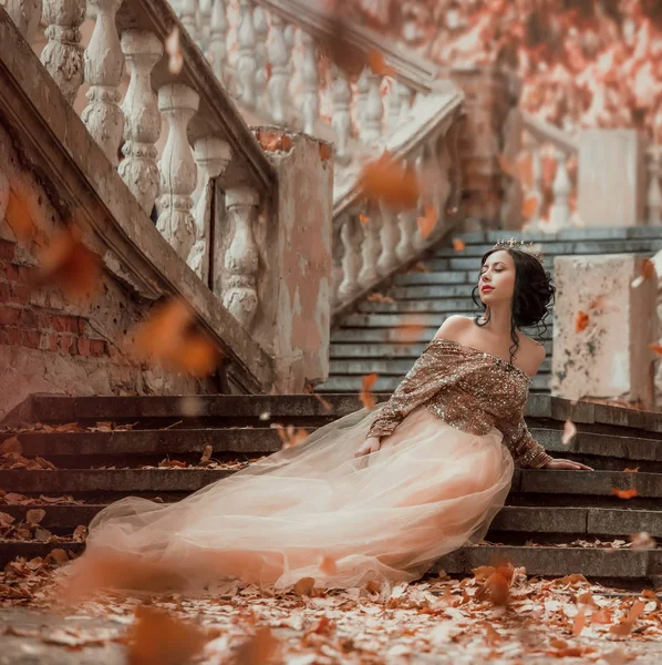 Affascinante ragazza bruna dai capelli scuri seduto da solo sui gradini di pietra di un incredibile castello reale, grazioso mago in un lungo elegante vestito d'oro che gioca con le foglie autunnali — Foto Stock