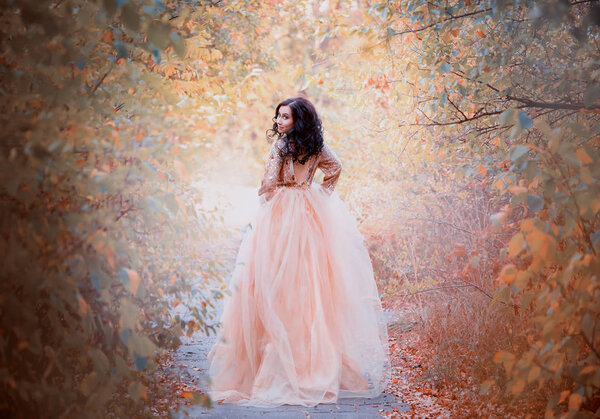 Charming brunette with dark hair girl runs away from the ball, the princess in a luxurious royal gold delightful dress walks along the forest path and turns around with a sweet smile.