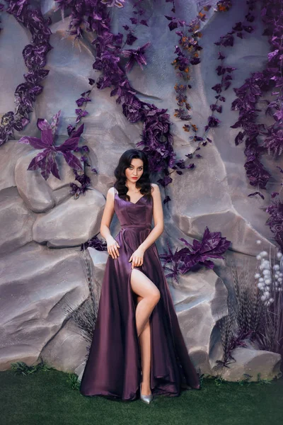 Sexy attractive lady with dark curled hair against stone wall with unusual magic plants, gorgeous charming countess in long, luxurious luxurious expensive purple satin dress, shows bare leg in cut — Stock Photo, Image