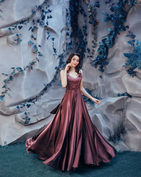 Charming fairy of flowers and flora stands near stone wall playfully poses for the camera in long magical elegant amazing marsala dress, attractive model like a doll with dark hair in a cold wave — Stock Photo, Image