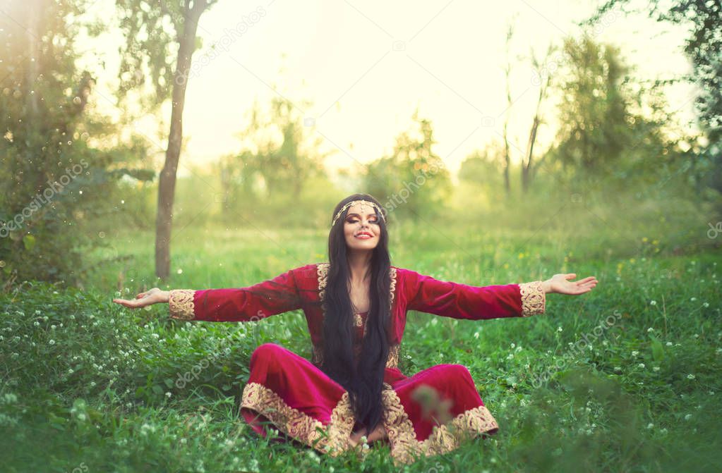 charming brunette girl in long burgundy maroon dress on grass in summer glade, wonderful Valida rests, meditates, enjoys sun, time of the Ottoman Empire, positive emotions, gold jewelry and tiara