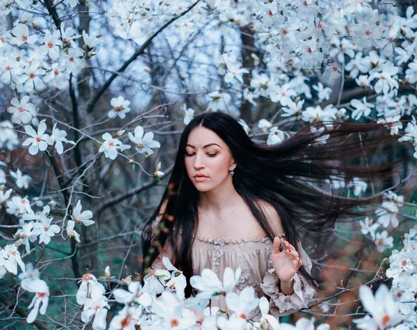 Wunderbare attraktive dunkelhaarige Dame mit geschlossenen Augen steht im Garten blühender Magnolien. Haare fliegen mit dem Wind, erstaunliche Foto in kalten Schattierungen von weißem Filter, Kunstbearbeitung Fotos — Stockfoto