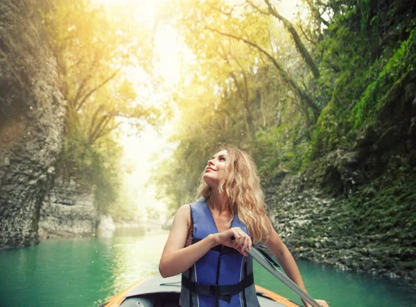El río azul de la montaña reluce en sol, encantadora muchacha sonriente feliz con pelo rubio exuberante se sienta en barco en el lago en Georgia, naturaleza Martvili Canyon, rafting como ocio activo para los turistas, sol brillante — Foto de Stock