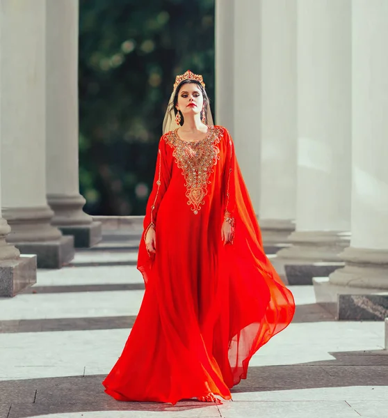 Prachtige Turkse keizerin shakherezad luxe rode jurk met mouwen, lange trein, goud vliegen. loopt soepel tussen grote kolommen met kroon, oorbellen en heldere dieprode lippenstift. kunst sexy foto — Stockfoto