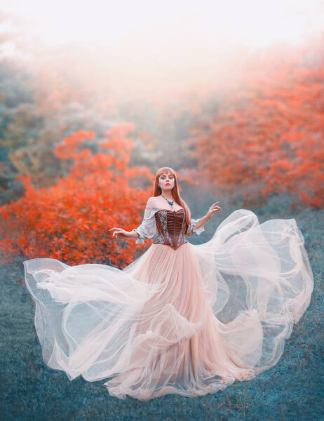 Charming attractive girl with long flying waving peach light vintage dress stands alone in forest, innocent lady looks into camera, gorgeous princess with red hair and magic necklace around her neck.