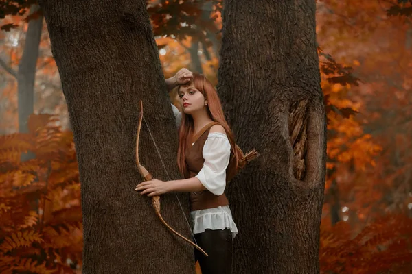 Misteriosa menina com cabelos vermelhos brilhantes entre as árvores em florestas de outono, encantador caçador com arco e flechas atrás de suas costas, elfo arqueiro atenciosamente observando animais da floresta, kitsune em forma humana — Fotografia de Stock