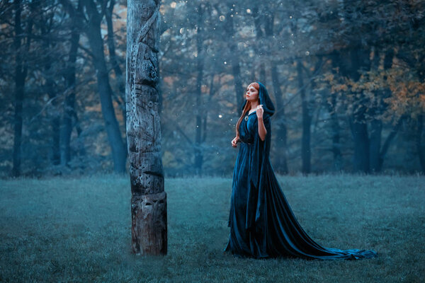Princess with red long hair dressed in blue expensive velvet royal cloak-dress, girl got lost in dark foggy forest, art photo, red-haired witch goes to totem monument in moonlight, pagan worship idol.
