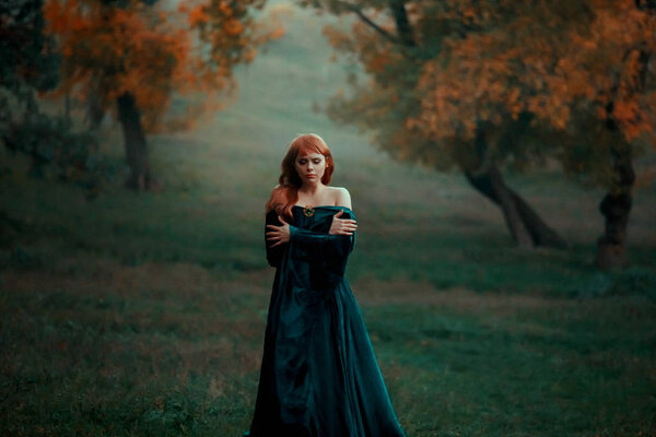 Lonely sad girl walks alone in terrible dark dangerous forest in long green emerald dress and raincoat with open shoulders, princess got lost and froze, young witch with red hair looks for path home.