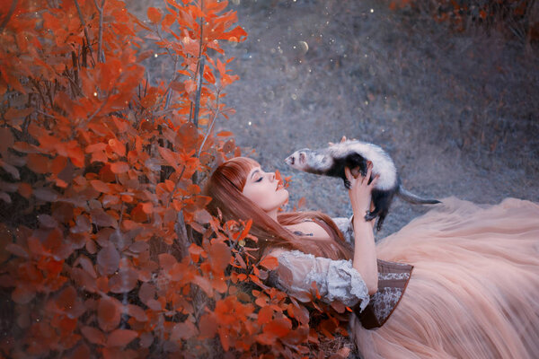 charming beauty lies on green grass in forest, princess in long, gorgeous light dress plays with a ferret as if she is a fairy-tale animal red-haired girl with closed hair under red autumn leaves