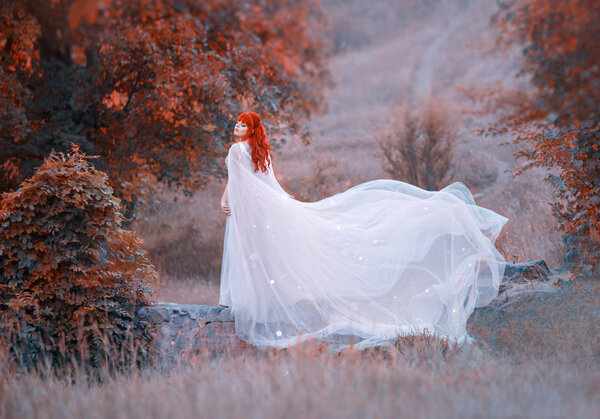 Charming girl with bright orange red curly hair on stone bridge in autumn forest, attractive elf princess in image of bride, wind waves white train of flying silver dress with sunlight, winter queen.