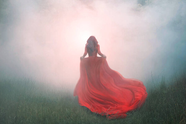 Gorgeous amazing wonderful scarlet red dress with a long flying waving train, a mysterious girl with red curly hair runs away into a thick white mist and forest haze over the green summer grass.