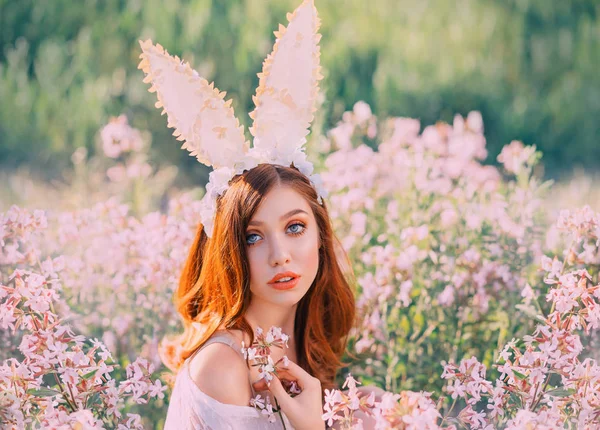 Girl easter bunny with creative ears on the hoop. Portrait of a young, red-haired woman with big beautiful eyes and lips. Rabbit sits in a flowery pink field. Gentle make-up natural in warm colors. — Stock Photo, Image