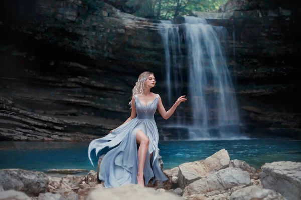 Una ragazza bionda riccia in un lussuoso abito blu siede su pietre bianche sullo sfondo di un paesaggio favoloso. Fiume Sirena vicino al lago con una cascata. Fotografia d'arte — Foto Stock