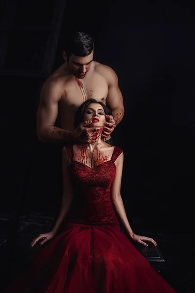 Bloody love story with vampire, girl and guy in dark room with scarlet blood on her arms, chest and face, scary stories of the game with Satan, sexy lady in red long dress and man holding her head — Stockfoto