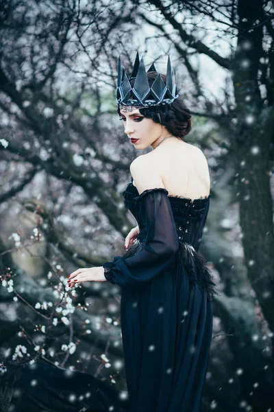 Grim dame met bleke huid en donker haar naast bloeiende boom, heks verandert met de zwarte kraai met lange luxe jurk en open rug en schouders, gotische beeld en make-up, koude metalen kroon en juwelen — Stockfoto