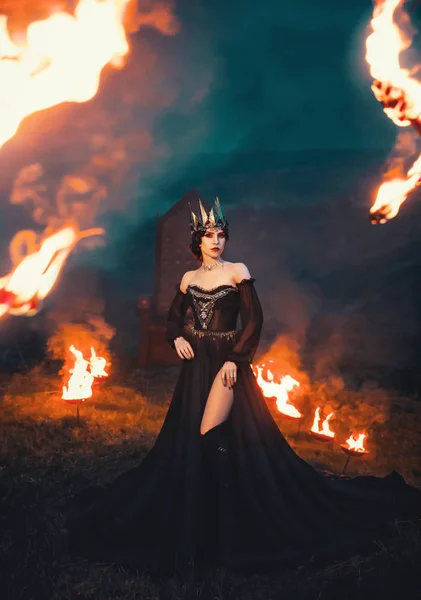 Σκοτεινή Βασίλισσα της νύχτας, μεγαλοπρεπή ιέρεια σε μακρύ μαύρο φόρεμα με γυμνούς τους ώμους, το κοράκι μάγισσα σε φλόγα, ισχυρό διάβολος με ωχρό δέρμα και μέταλλο κρύο στέμμα στο κεφάλι της, ερωμένη και φωτιά θεά — Φωτογραφία Αρχείου