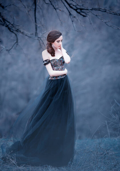 Gorgeous attractive brunette, lady in a long black dress with bare open arms and shoulders, the girl alone in the autumn cold forest, the weather in November is a dark princess in the form of raven.