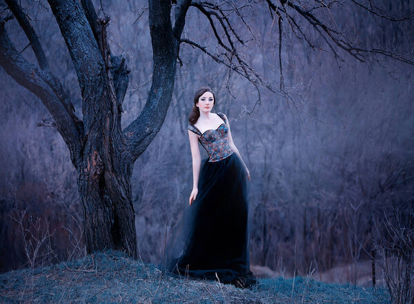 Gorgeous attractive brunette, lady in a long black dress with bare open arms and shoulders, the girl alone in the autumn cold forest, the weather in November is a dark princess in the form of raven.
