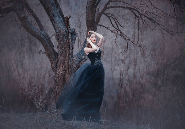 Amazing tender brunette, lady in a long black dress with bare open arms and shoulders, the girl alone in the autumn cold forest, the weather in November is a dark princess in the form of crow.