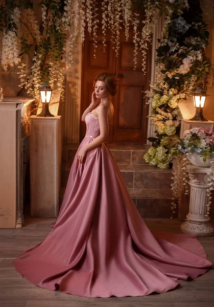 Gorgeous tender red-haired girl in a long, chic satin pink gorgeous dress with salted shoulders, a pretty attractive fine princess walks alone in a flower yard, nude makeup, glare of lanterns — Stock Photo, Image