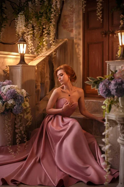 Gorgeous tender red-haired girl in a long, chic satin pink gorgeous dress with salted shoulders, a pretty attractive fine princess walks alone in a flower yard, nude makeup, glare of lanterns — Stock Photo, Image