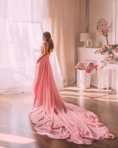 Imagem da primavera, senhora doce e gentil em vestido rosa lindo longo olhando diretamente para a câmera,, uma menina como uma flor de rosa, fica com ela de volta para a câmera perto de grande janela, cores nuas — Fotografia de Stock