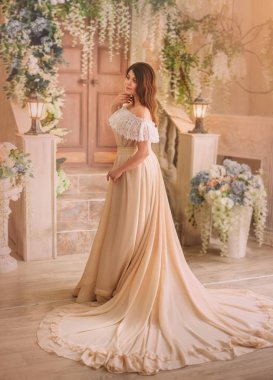 Esmer elbiseli bir kız bir krem vintage Açık omuz ile ve uzun güzel bir tren. Model bir çiçekli avlu karşı poz. Nazik doğal makyaj. Ortaçağ Prenses