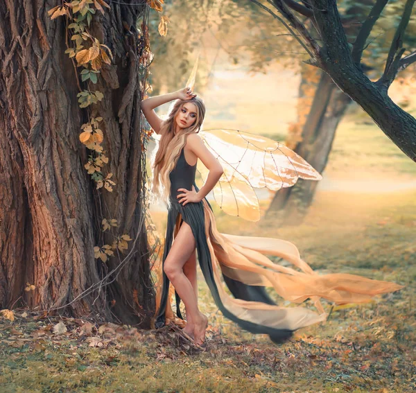 Gentle girl with blond hair poses for camera in forest, wonderful fairy-tale fairy with transparent wings in long green dress with flying split train, mythical creature, spirit. free spase for text — Stock Photo, Image
