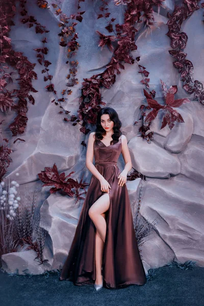 Sexy attractive lady with dark curled hair against stone wall with unusual magic plants, gorgeous charming countess in long, luxurious expensive purple satin dress, shows bare leg in cut — Stock Photo, Image