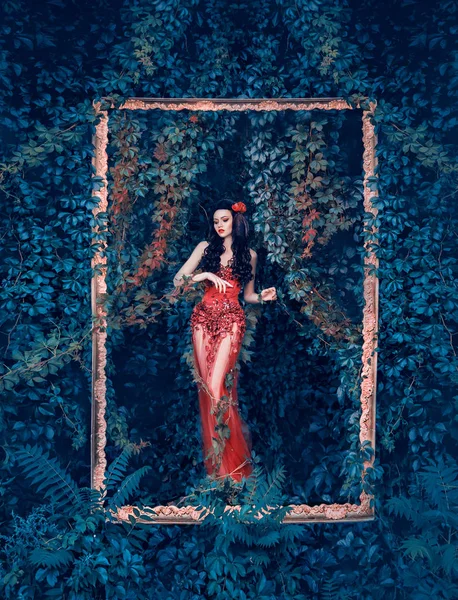 Misteriosa diosa del bosque y la naturaleza sale de su jardín en un elegante vestido rojo con un largo tren transparente y decoración floral de diamantes. chica atrapada en el arte, morena está atada por las vides de plantas —  Fotos de Stock